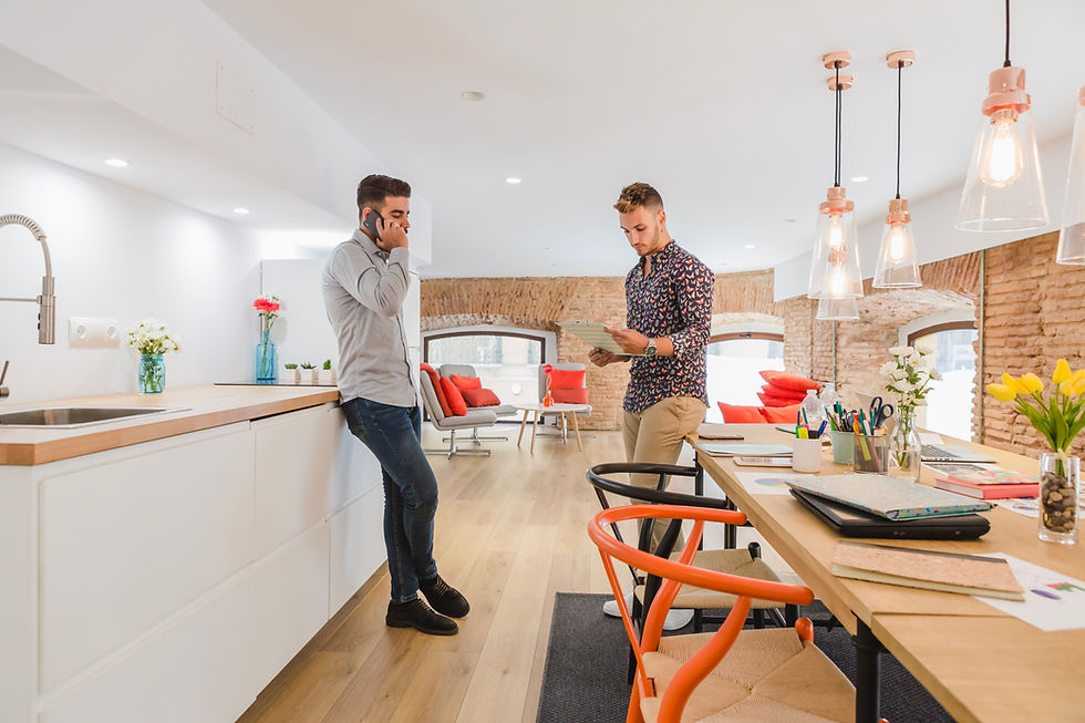 Two men in a stylish loft office; one on the phone, the other reading documents. Bright decor with orange chairs and flowers on tables.