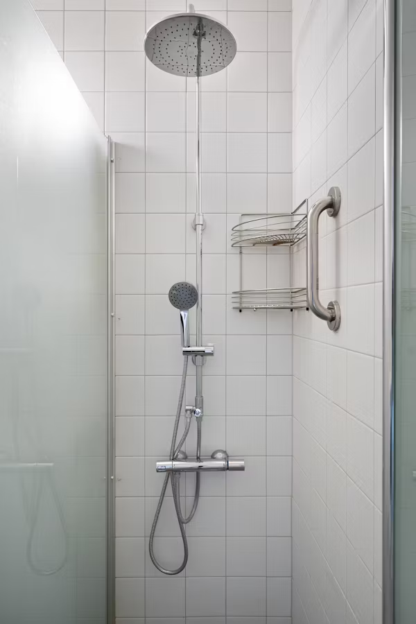 Modern shower with dual heads and metal rail on tiled wall. Glass door and shelf are present, creating a clean, minimalist look.