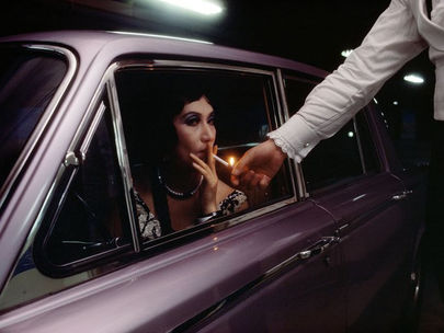 Photo : René Burri. Un homme en livrée allume la cigarette d'une femme au volant de sa voiture, qui regarde l'objectif.