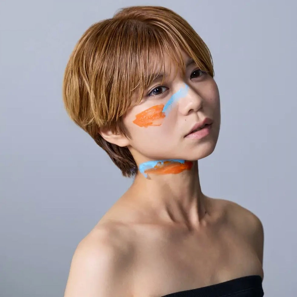 青い背景の前で、金髪の短髪の人物が肩を出してポーズ。頬と首に青とオレンジのペイントがあり、静かな表情。