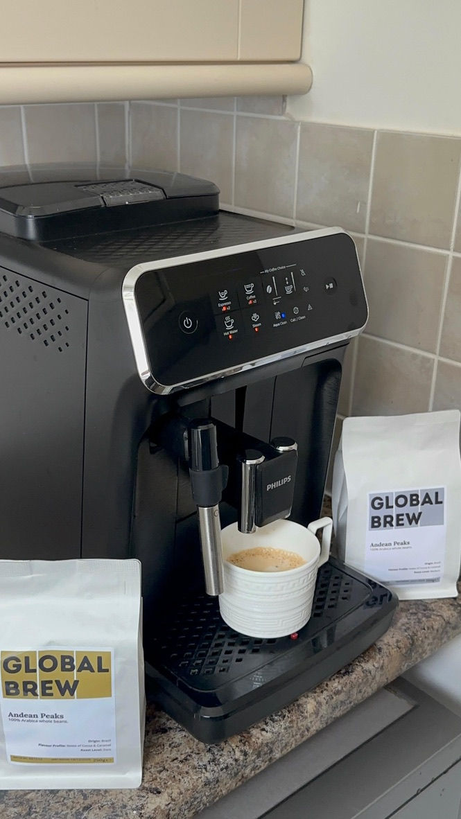 Coffee machine brewing into a white mug on a kitchen counter. Two "Global Brew" coffee bags labeled "Andean Peaks" are beside it.