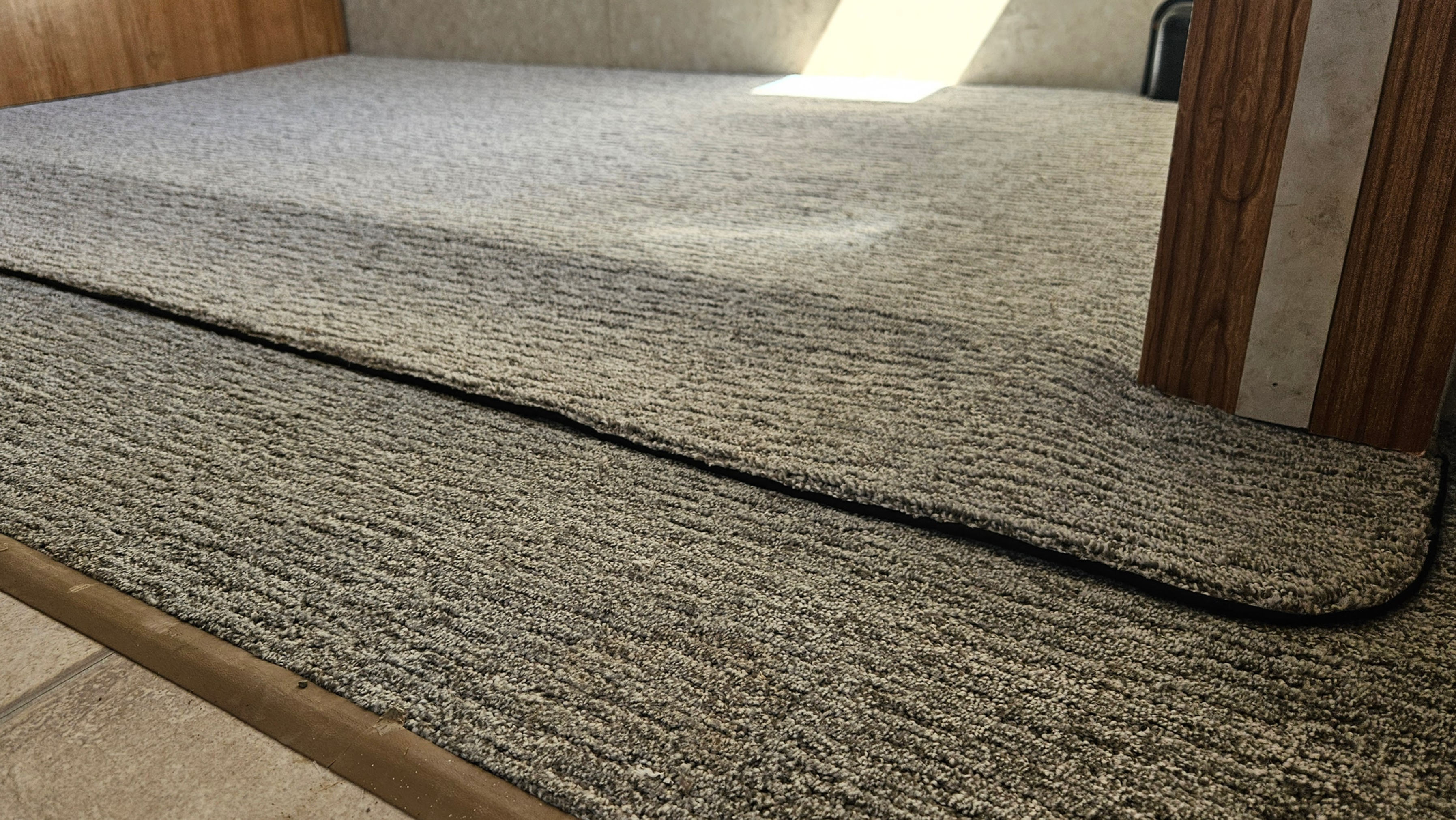 Close-up of newly installed grey carpet in an RV slide-out, featuring a neatly sewn fabric binding along the edge for a clean, professional finish. The carpet transitions smoothly from the main floor to the elevated slide platform.