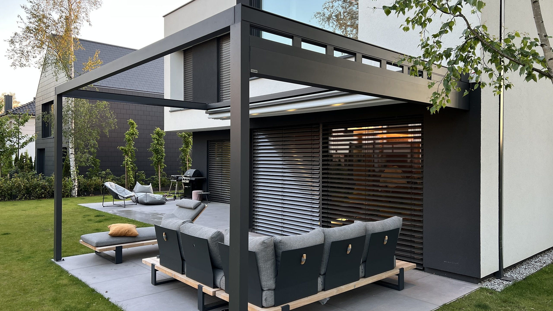 Freestanding anthracite retractable pergola with the fabric roof folded back over a terrace sofa beside a modern house