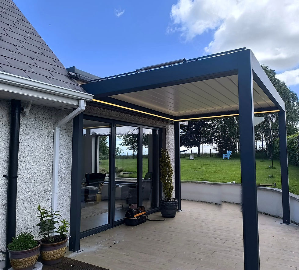 Anthracite louvred pergola with four posts on a curved patio beside a white house, warm LED strip glowing under the roof, garden and trees behind