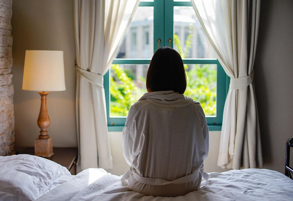 Person sitting quietly on a bed looking out a window, representing calm, rest, and relief from tension.