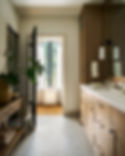 Spacious bathroom with wooden cabinets, a beige tiled floor, and a window with sheer curtains. A plant and towels sit on a wooden table. on HomeFindss by STEVE & FILIP DESIGN