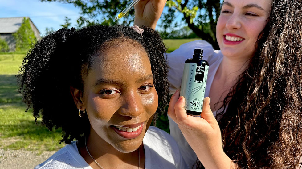 A smiling woman applying hair growth oil to another woman’s scalp outdoors using a dropper, with natural scenery in the background.