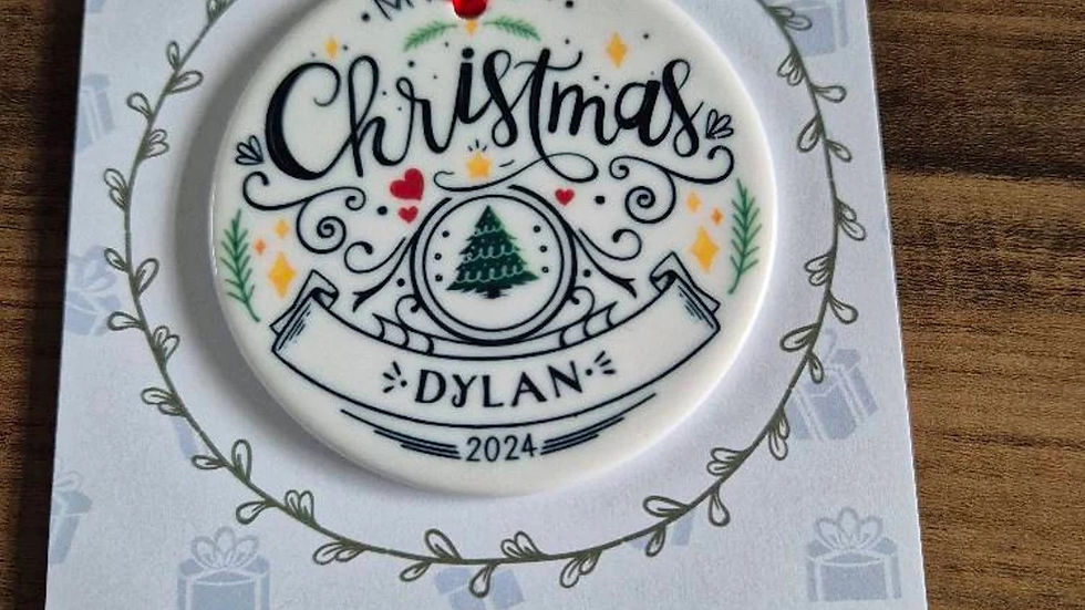 White ornament with "My 1st Christmas Dylan 2024" hangs by red ribbon. Background features gift icons and company info.