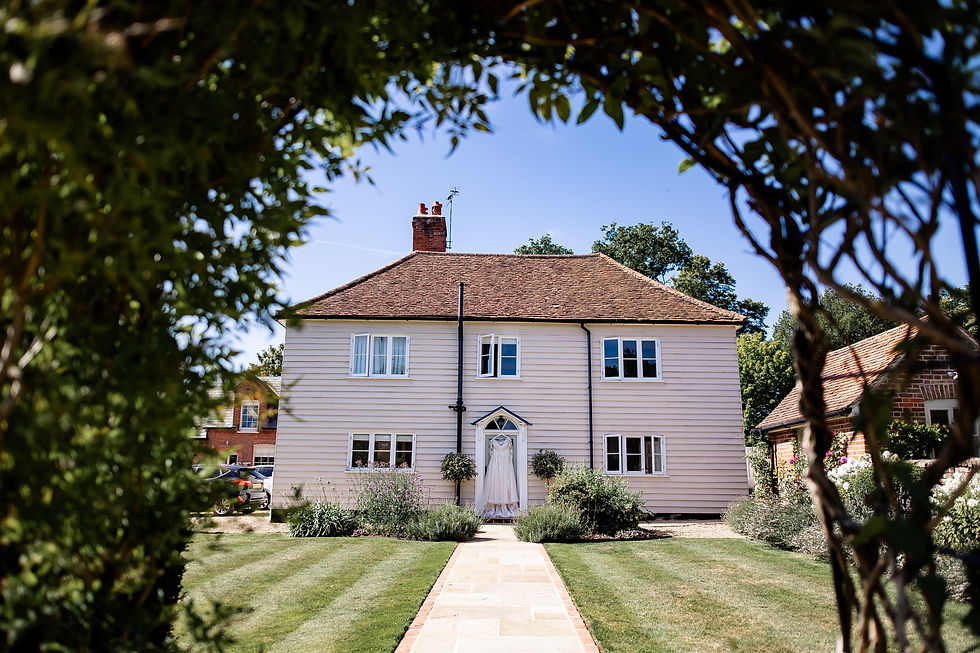 The house at Blake Hall with brown roof framed by greenery, wedding dress hanging on front door. Sunny day, blue sky, manicured lawn, cozy mood.