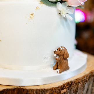 White wedding cake with gold accents and flowers, set on a wood slab. A small brown fondant dog appears to be eating crumbs at the base at Wellington Wood wedding venue in Norfolk