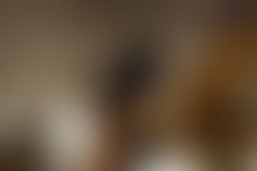 Fotografia em cores realizada a partir do interior da exposição. Em primeiro plano, à direita, uma escultura em madeira clara aparece desfocada e ocupa parte significativa da imagem, revelando volumes arredondados, figuras humanas e formas vegetais entrelaçadas. Ao centro, um homem é visto de costas caminhando entre as obras, com as mãos cruzadas atrás do corpo segurando um catálogo. À esquerda, outra escultura repousa sobre pedestal branco, expandindo-se horizontalmente em raízes e prolongamentos orgânicos. Ao fundo, uma escultura vertical de grande porte ocupa posição central, cercada por outras peças distribuídas pelo espaço. Uma abertura em arco revela parcialmente outro ambiente da galeria. A composição utiliza diferentes planos de foco para destacar a experiência do percurso expositivo, sugerindo uma imersão gradual entre as esculturas e o espaço arquitetônico.