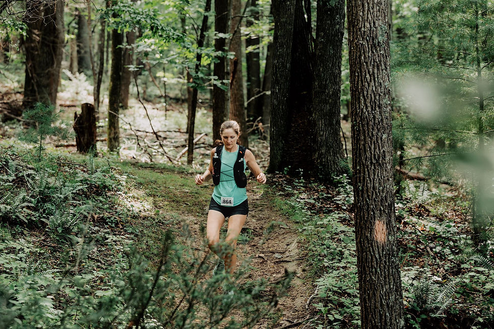Female trail runner wearing bib 864 runs along a wooded forest path, focused, surrounded by green trees and ferns.