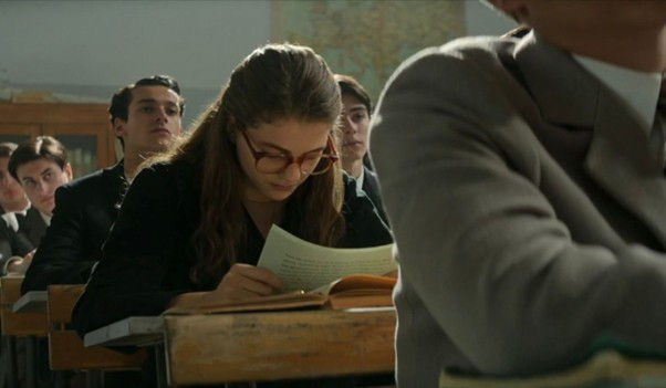 A imagem mostra alunos em uma sala de aula, sentados e olhando para frente. Ao centro, uma das personagens principais da série, está olhada para baixo e lendo algo escrito em folhas de papel.