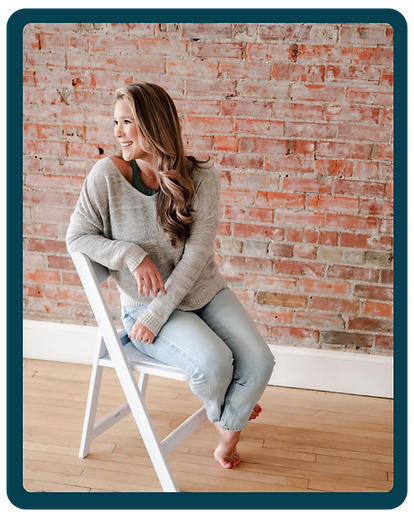 A woman sitting on a white chair in front of a brick wall smiling and looking over her right shoulder.