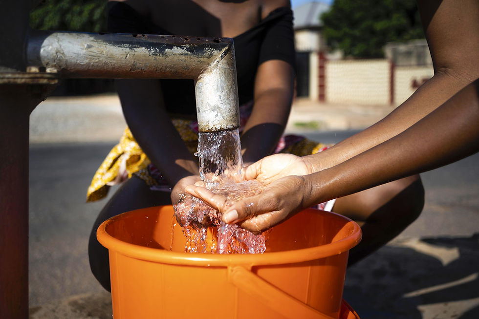 Hands collect water from a pipe into an orange bucket. Bright sunlight, outdoor setting. A person is visible in the background.