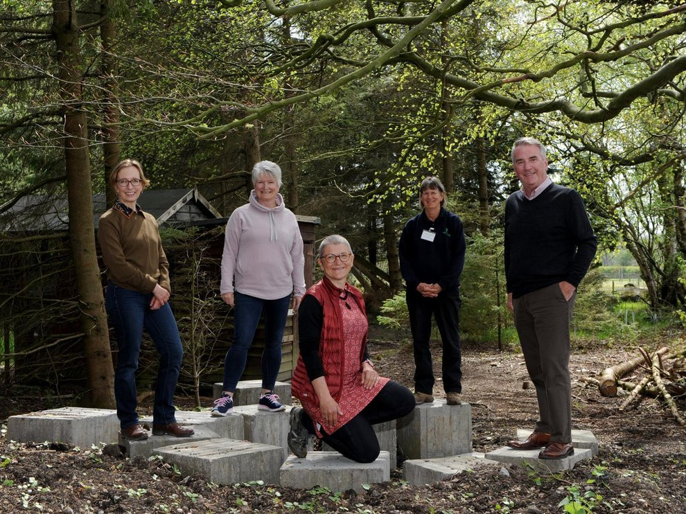 Five people surrounded by trees. Four are standing, one is kneeling in the centre. All are smiling at the camera.
