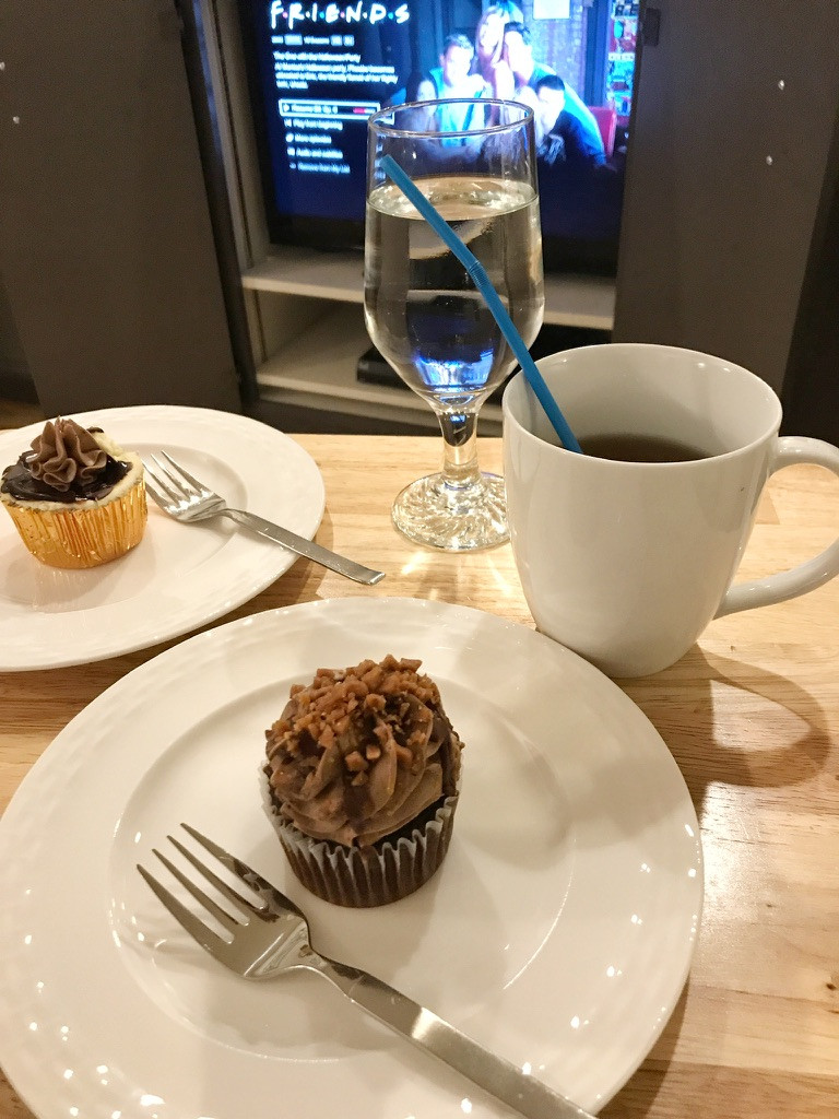 ID: A tray of two cupcakes and two drinks with the TV show Friends playing in the background.