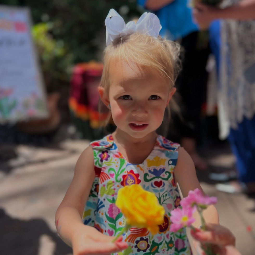 Smiling young girl holding colorful flowers at a bloom bar celebration, wearing a floral dress with a white bow in her hair.