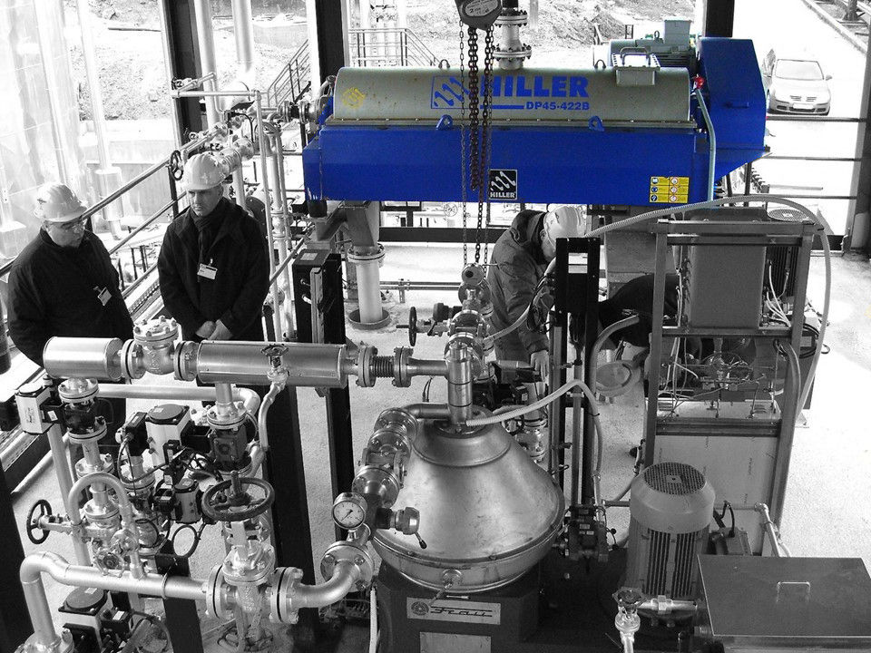 Two workers examine a dewatering centrifuge setup in a processing area.