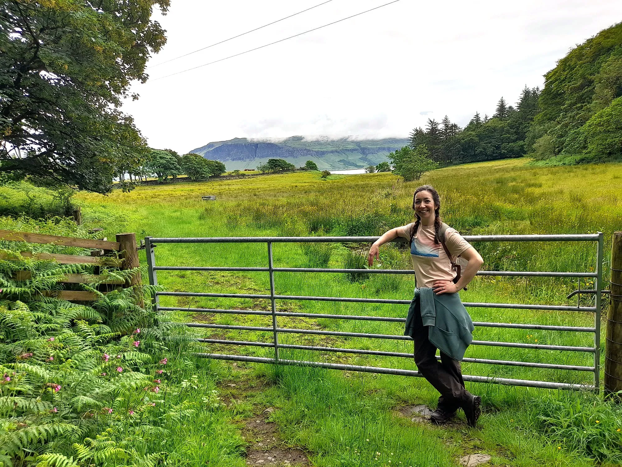 A woman standing leaning on a farm gate wearing a pink t-shirt and a green hoodie tied around her waist, smiling. Behind her is a lush field, and a view of water and hills beyond.