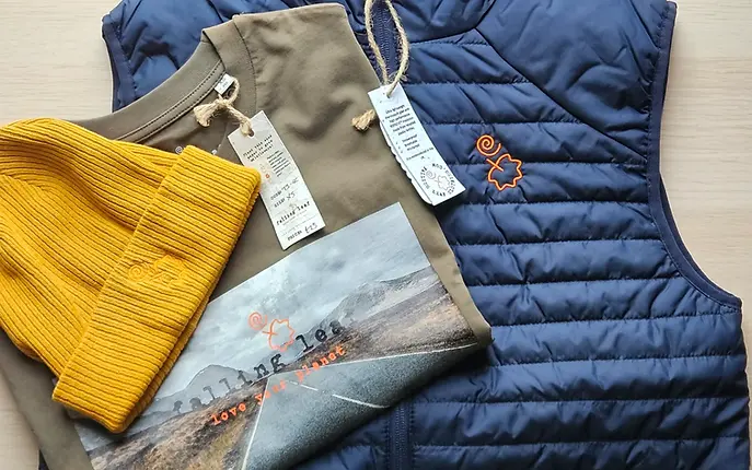 Blue gilet on a table, lying under a folded khaki t-shirt with the road to Glencoe printed on it, and a mustard coloured beanie on top of that