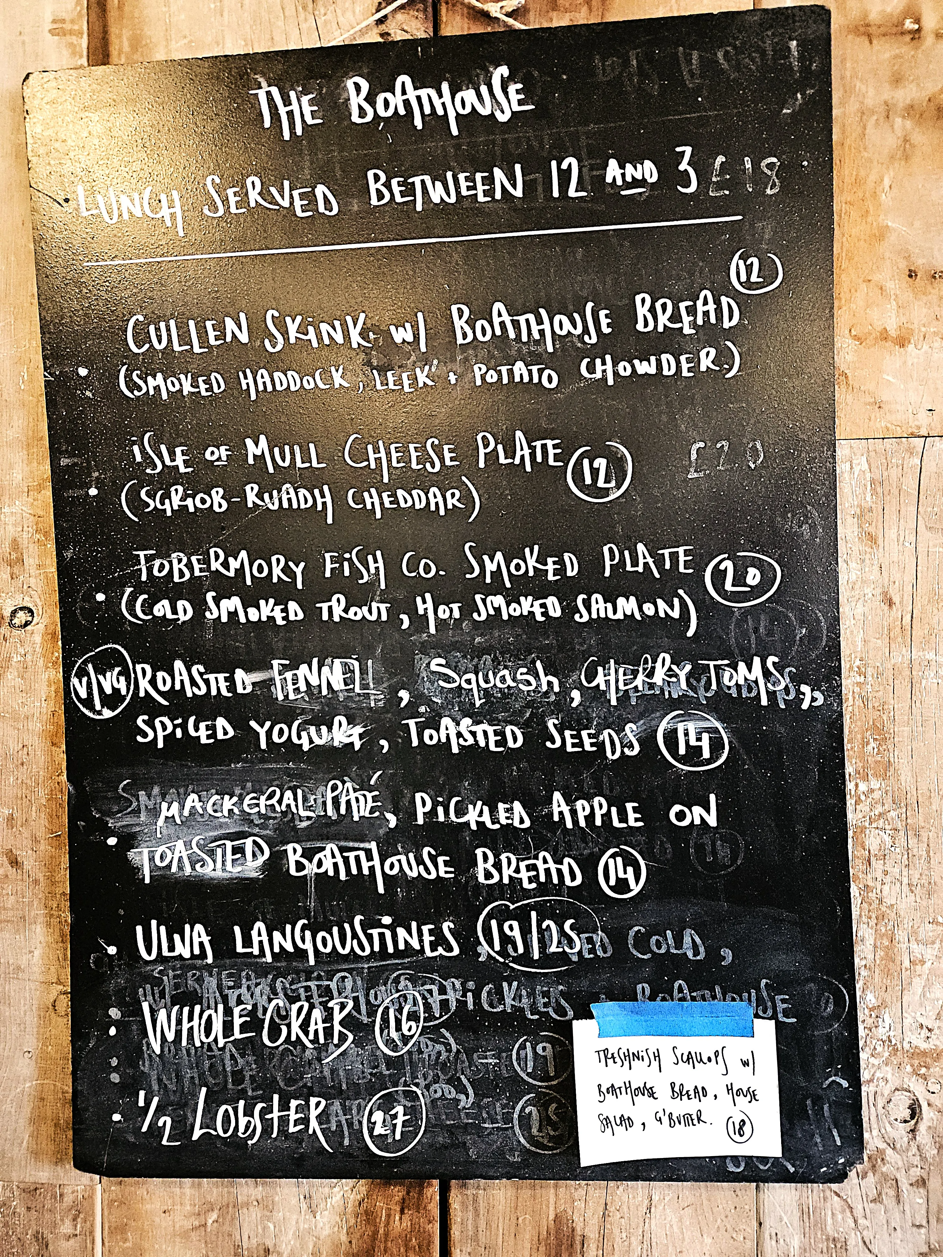 The menu board at The Boathouse - white chalk writing on a blackboard, stating lunch served between 12 and 3, then listing the options of cullen skink, mull cheese plate, smoked fish plate, roasted fennel and squash, mackerel pate, langoustines, whole crab, 1/2 lobster, and a paper note with the specials taped to it - Treshnish scallops.