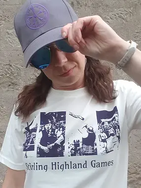 Woman wearing a grey cap and white t-shirt both with Stirling Highland Games embroidered/printed on them. The t-shirt also has images from a highland games - dancers, bagpipes, a caber and a footrace. She is holding the peak of the cap and wearing dark sunglasses