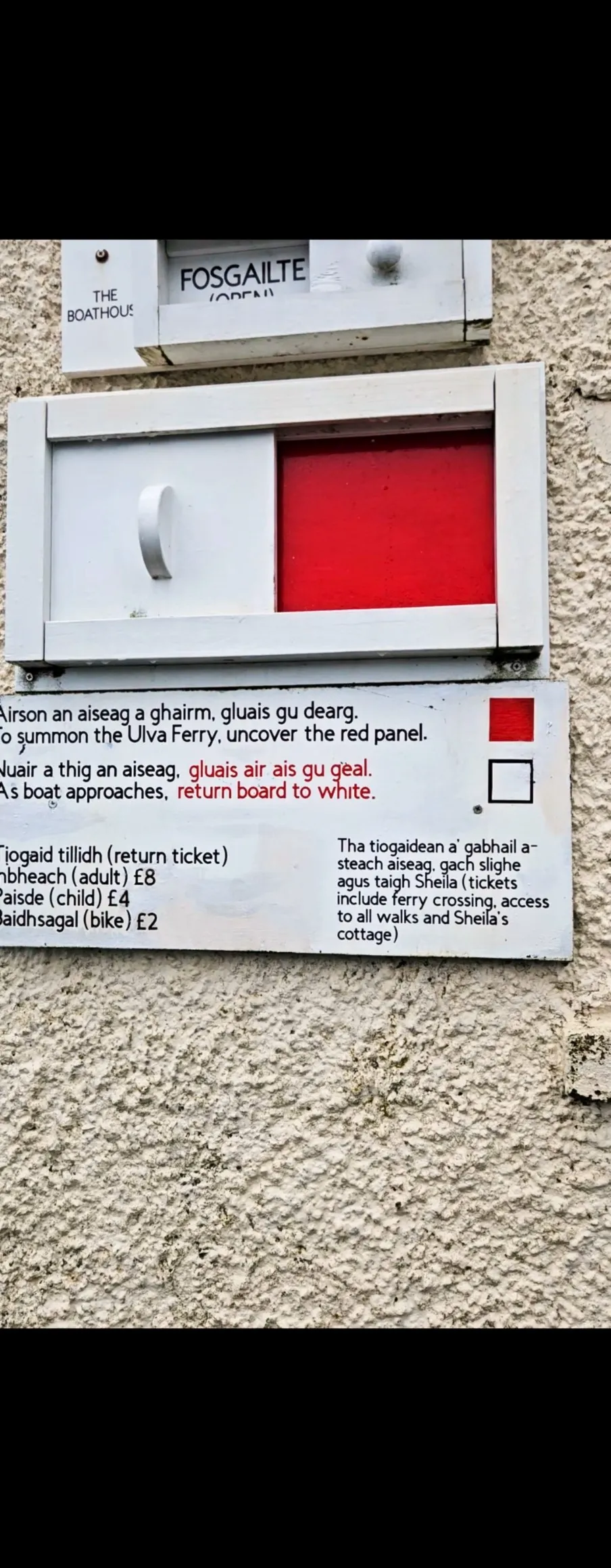A wooden frame mounted on a pebbledash wall, with a sliding hatch that allows the user to reveal a large red panel to hail the ferryman, with usage instructions and pricing below in English and Gaelic. A similar slider above shows that The Boathouse is open.