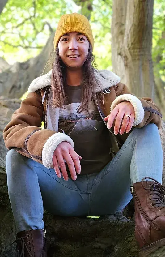 A woman crouched in a tree wearing the Glencoe t-shirt in khaki with a sheepskin jacket over it, light jeans, brown boots and a mustard coloured beanie