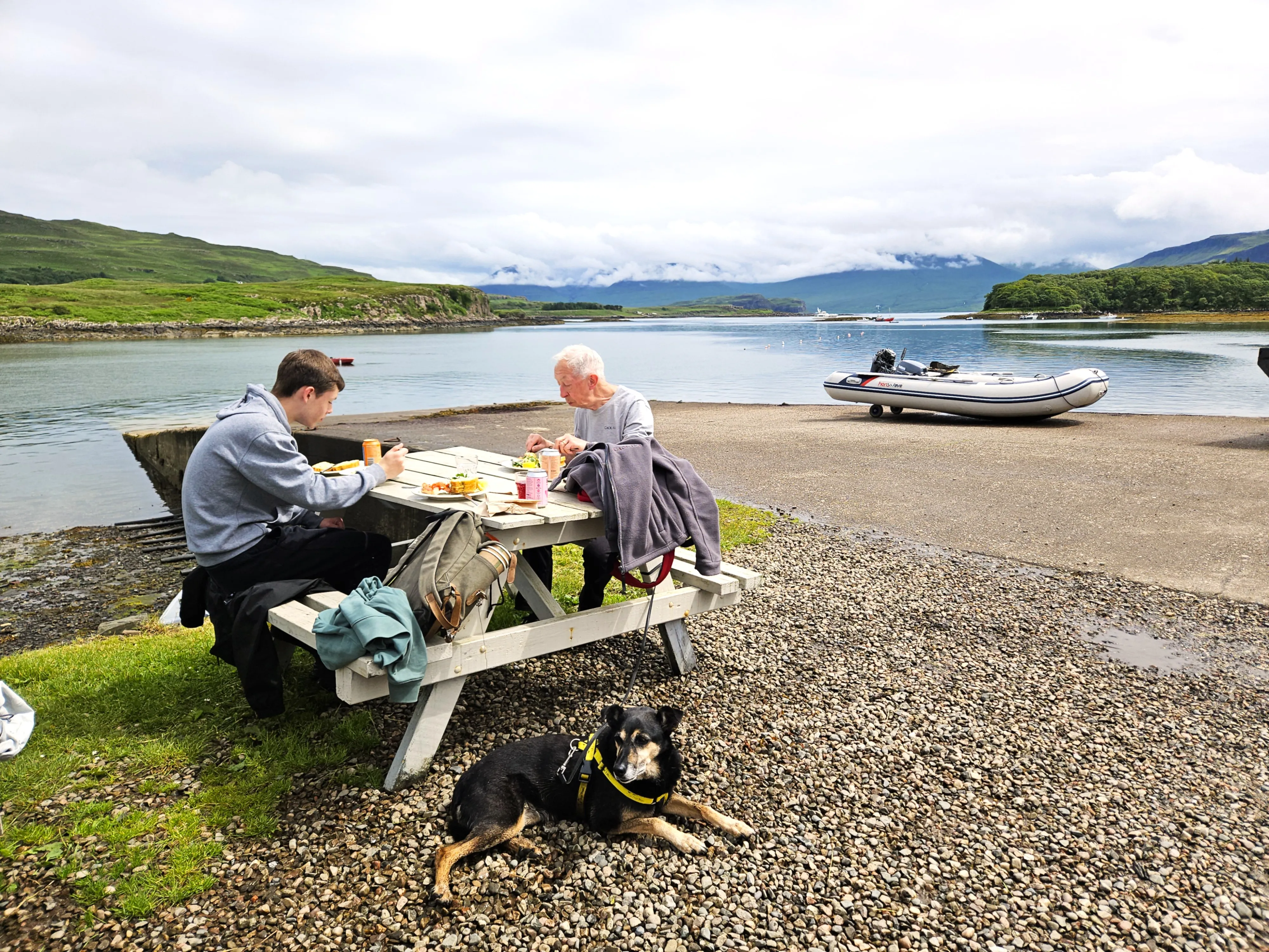 Two men sitting outside in the sunshine eating lunch at a picnic bench with a dog lying down in the foreground, and the ferry jetty and Mull in the background.