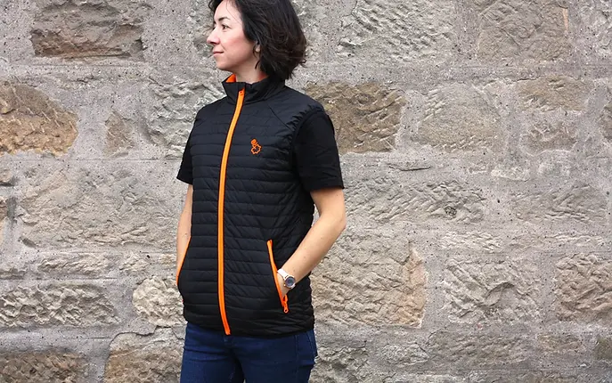 Woman wearing a black gilet with orange main zip and pocket zips, over a black t-shirt and dark jeans. Her hands are in the gilet pockets.
