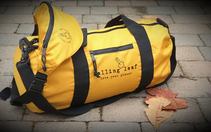 Mustard coloured duffel bag with black handles, with a matching coloured bum bag sitting on top of it, surrounded with auumn leaves