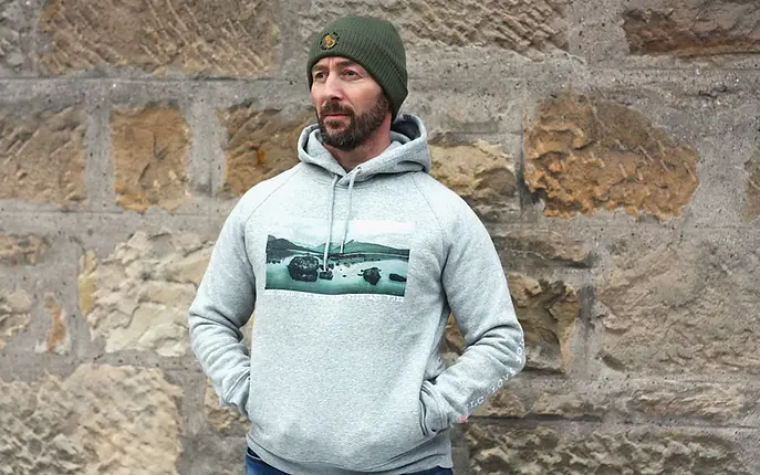 Man wearing a light grey hoodie with a loch print in black and green, with a green beanie, hands in the hoodie pockets.