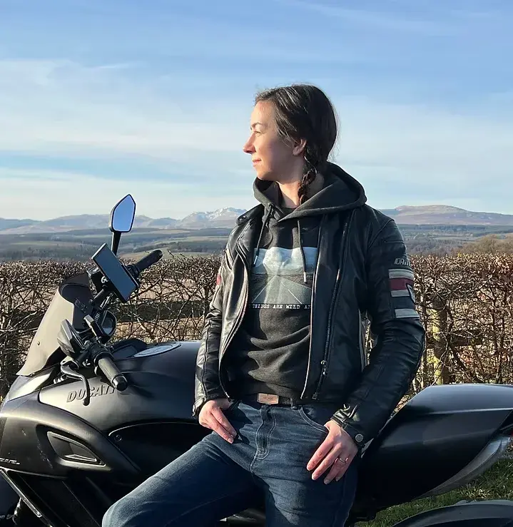 Falling Leaf founder Lindsay leaning against her motorbike (a Ducati called The Judge) wearing the Glencoe hoodie under a leather jacket, in front of a hilly view, with sunset lighting