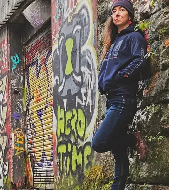 Falling Leaf founder Lindsay leaning on a brightly graffitied wall wearing the Duke hoodie and a grey beanie, dark jeans and brown boots