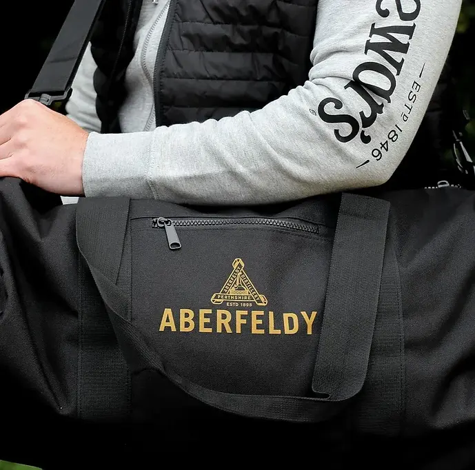 Close up of a black duffel bag with the Aberfeldy distillery logo in gold, held by someone wearing a light grey hoodie with Dewar's printed down the sleeve, under a black gilet