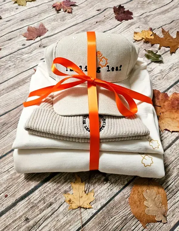 A folded bundle of clothing - natural coloured sweatshirt, t-shirt, beanie and cap tied with a bright orange ribbon surrounded withautumn leaves