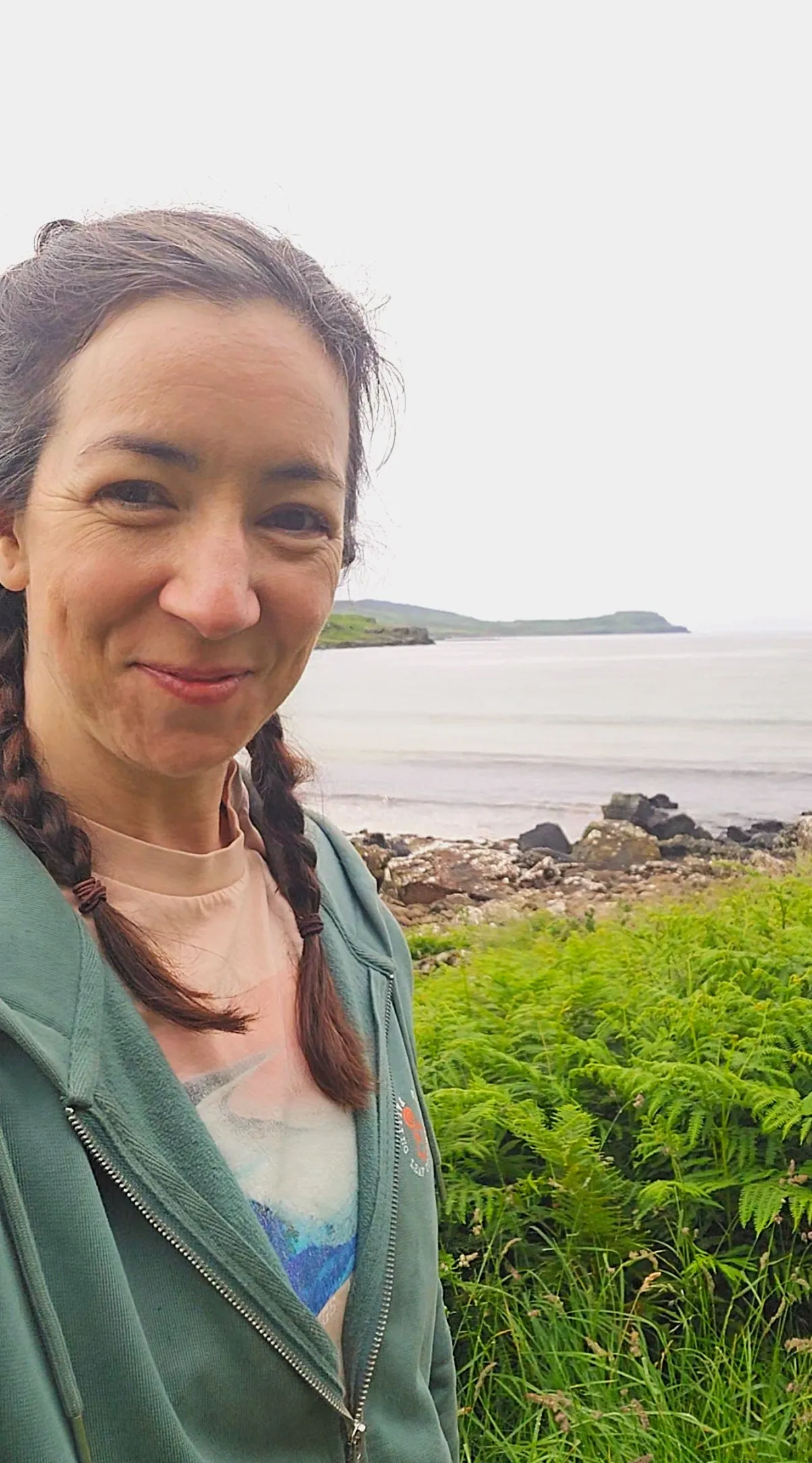 A woman with pigtails in a pink t-shirt and green hoodie in front of a fern-y, rocky beach on a hazy day.