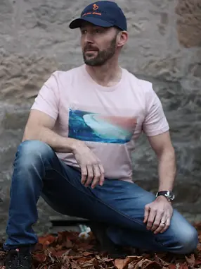 Man in a pink t-shirt with a blue white and pink print of a beach (Mill Bay in Orkney), blue jeans and a navy cap, crouching in autumn leaves