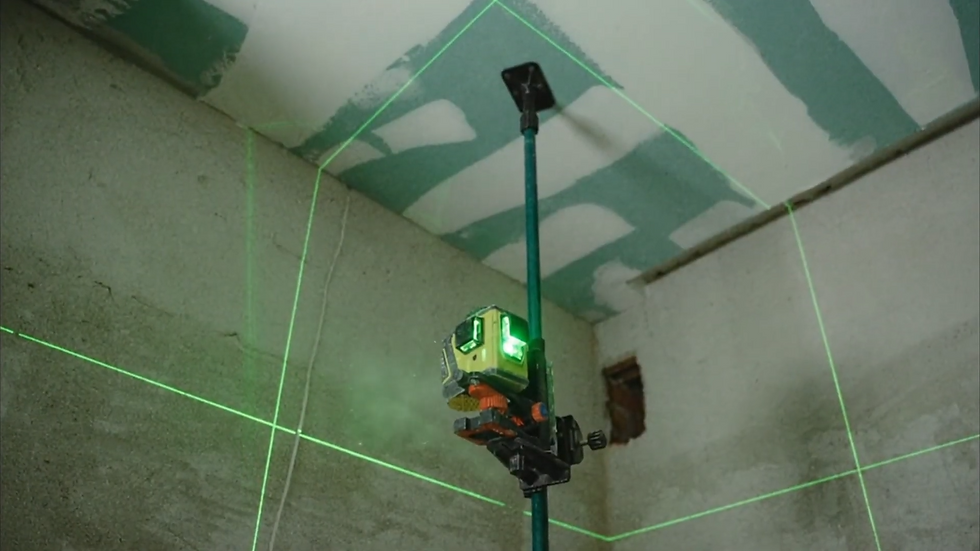 Laser level projecting green lines on a concrete ceiling in a construction setting, highlighting corners and edges. Dust is visible in the air.