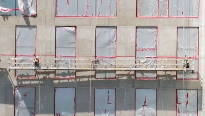 Workers on a suspended platform clean windows on a high-rise building. Windows are covered in protective plastic. The mood is industrious.