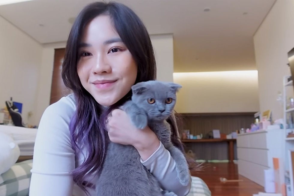 Perempuan berambut panjang dengan kaos putih menggendong British Shorthair abu-abu di dalam kamarnya.