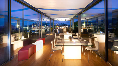 Toit-terrasse moderne avec bar, tables lumineuses, chaises design, vue nocturne.