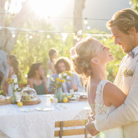 Bride and groom embrace at outdoor wedding with guests dining at a table. Warm sunlight and string lights create a romantic atmosphere.