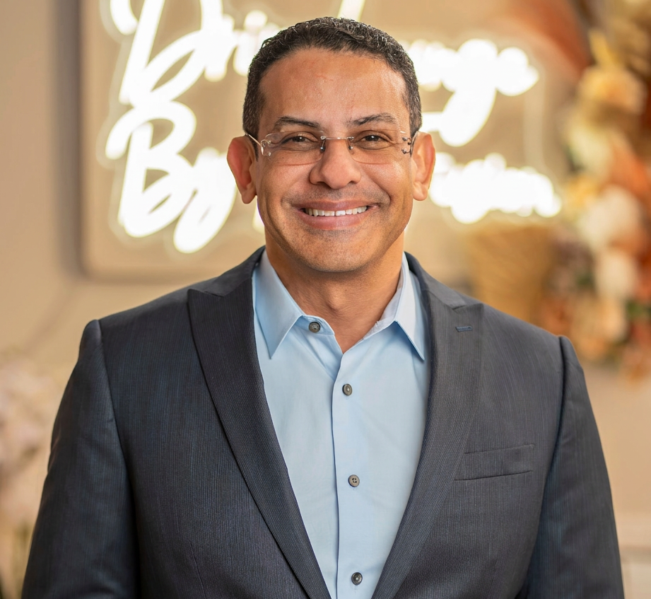 Studio photography of a smiling man in a suit and glasses. He appears confident and professional in a modern setting with subtle background decor.