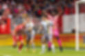 Daniel Jinadu in a bright pink football outfit with hands in the air, behind two teammates blocking an opponent from the goal during a match