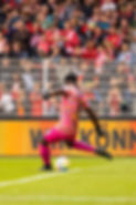 Daniel Jinadu in a bright pink football outfit, poised to strike the ball from the penalty area during a match, showcasing skill and focus