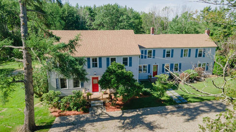 Large light-gray building with red doors, blue shutters, and surrounding green trees.