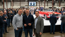 Three men posing at a live classic car auction with a red convertible on the auction block behind them