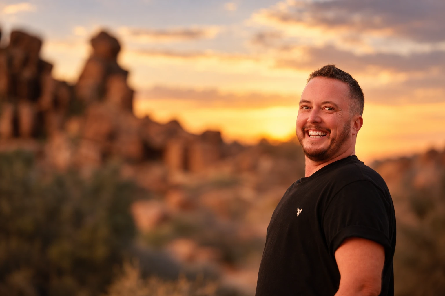 Ayce Kyptyn smiling in a black shirt against a warm Colorado sunset and rocky landscape
